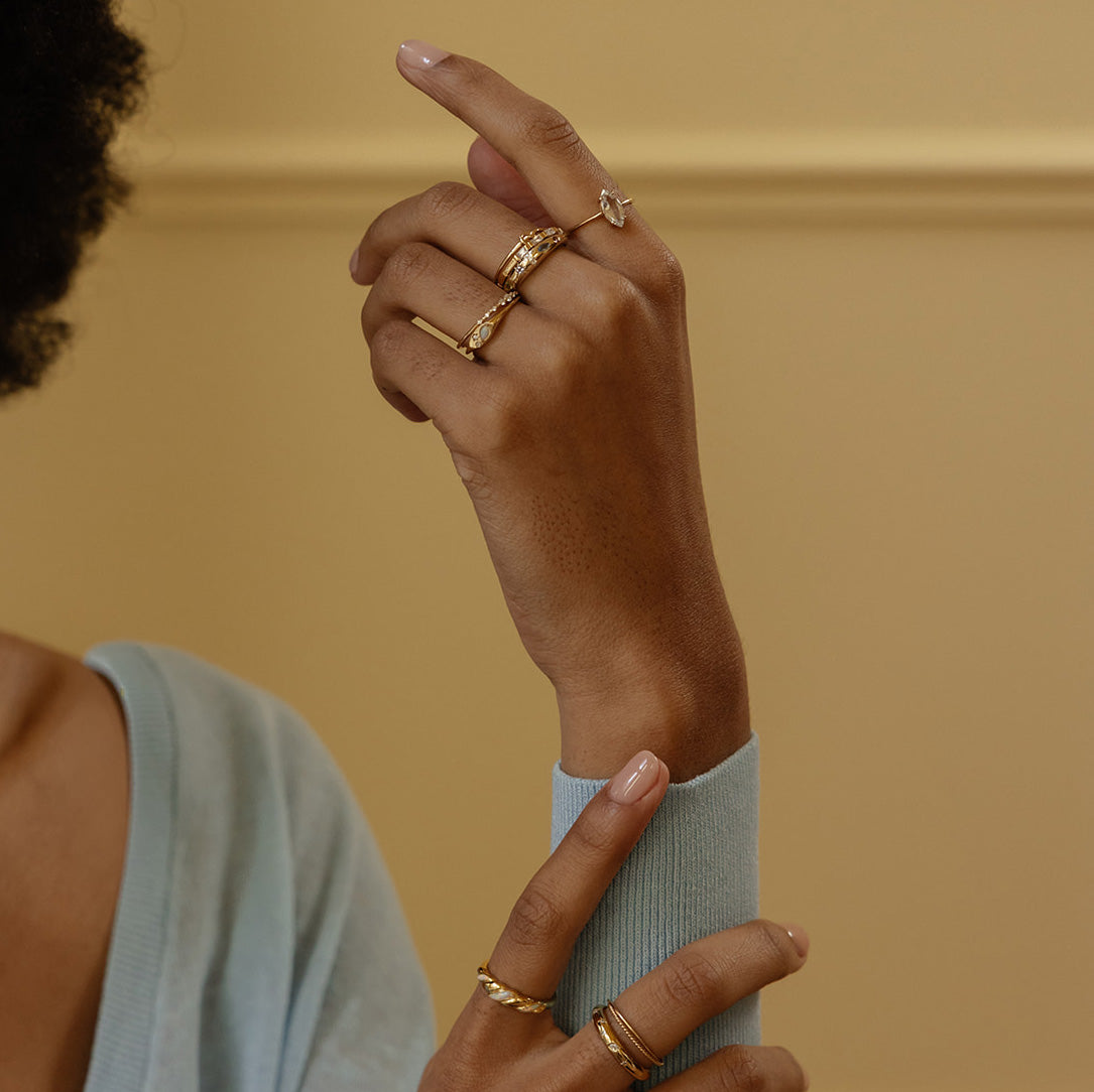 A person with light brown skin wears the Solid Gold Gemstone Marquise Ring in White Topaz on their finger, along with other gold rings, and a pale blue top. Their hands are gracefully posed against a beige background.