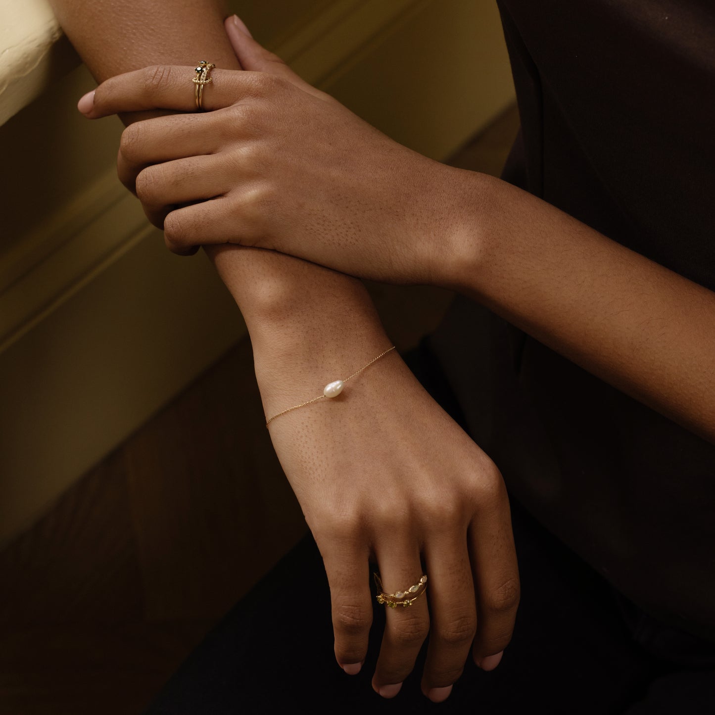 A person’s crossed arms display the Solid Gold Single Pearl Chain Bracelet and gold rings, set against a dark outfit background.