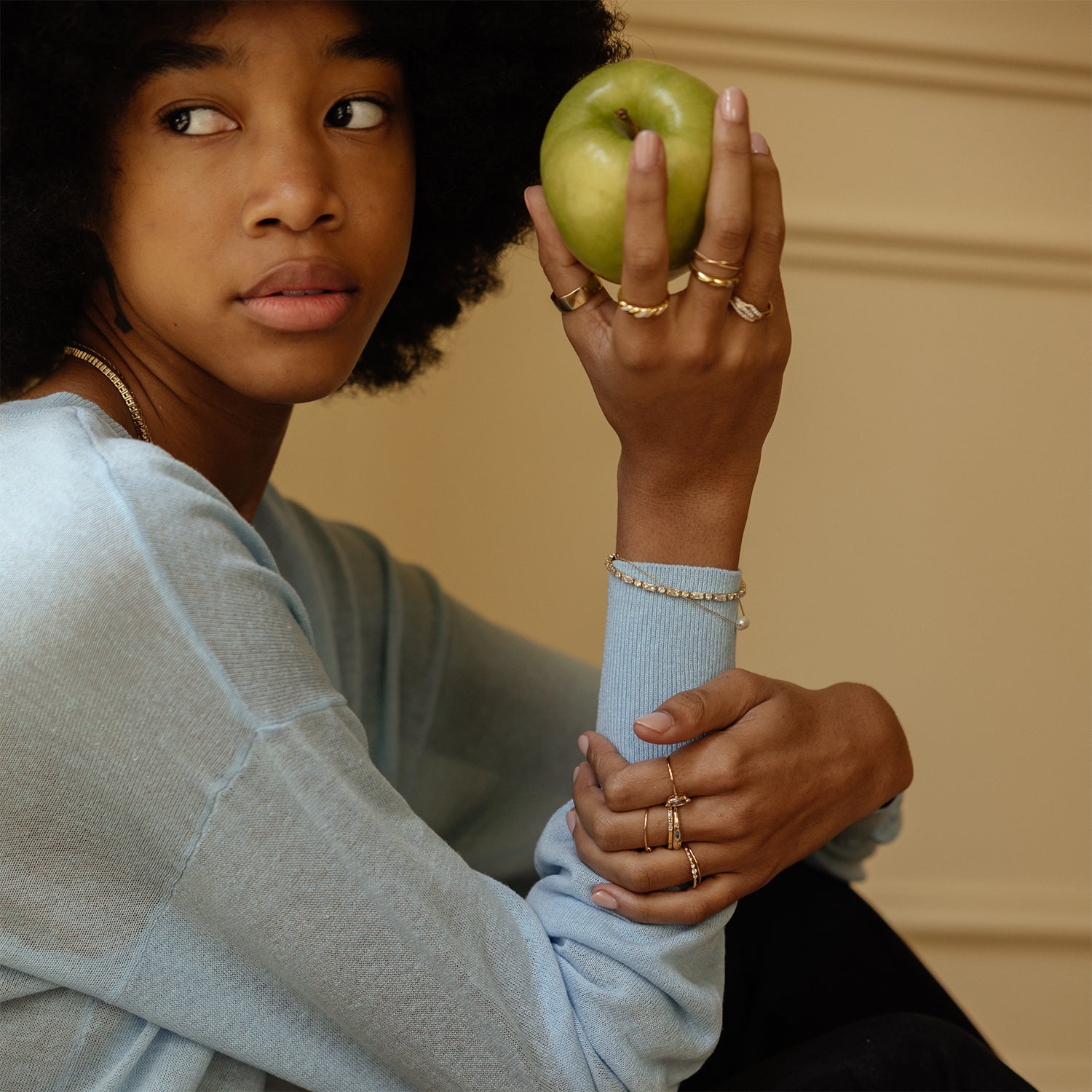 A person with an afro hairstyle in a light blue sweater holds a green apple near their face, looking to the side. They wear multiple gold rings and a sparkling Baguette Tennis Bracelet on both hands.