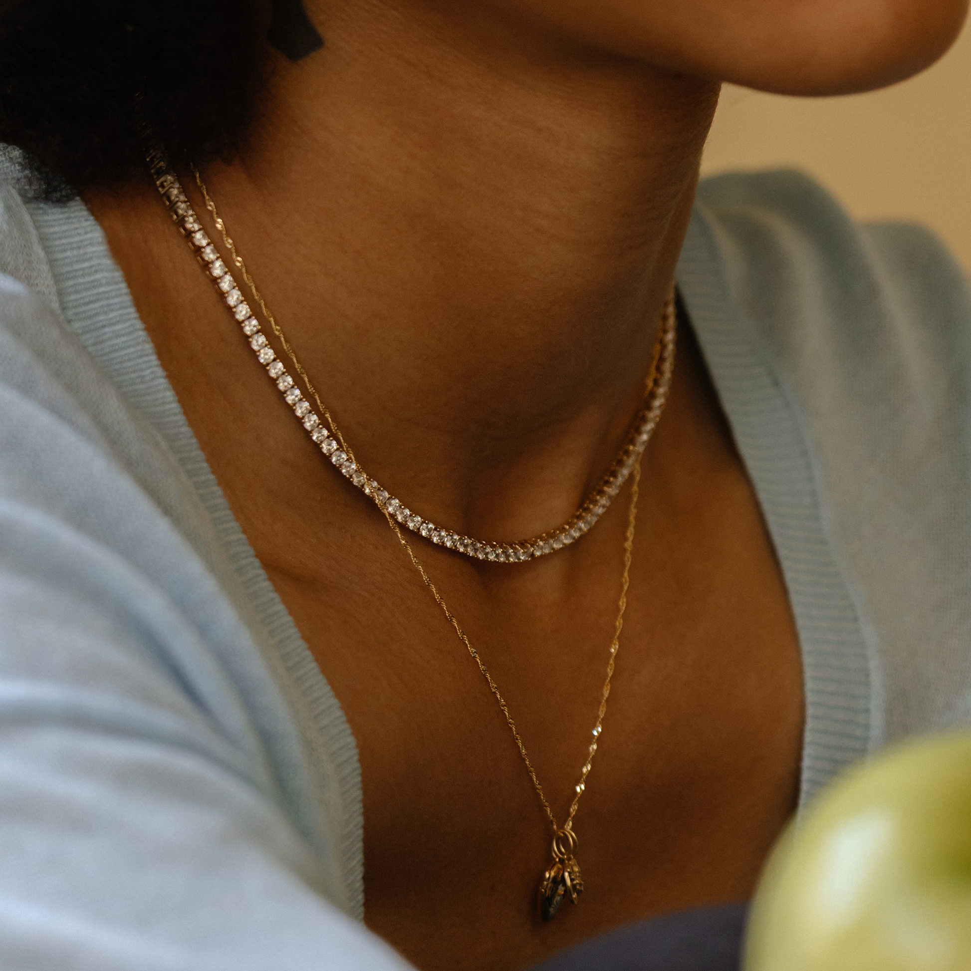 A close-up of a person wearing a 14k yellow gold plated chain with a small pendant and the sparkling Tennis Necklace, both paired with a light blue top.