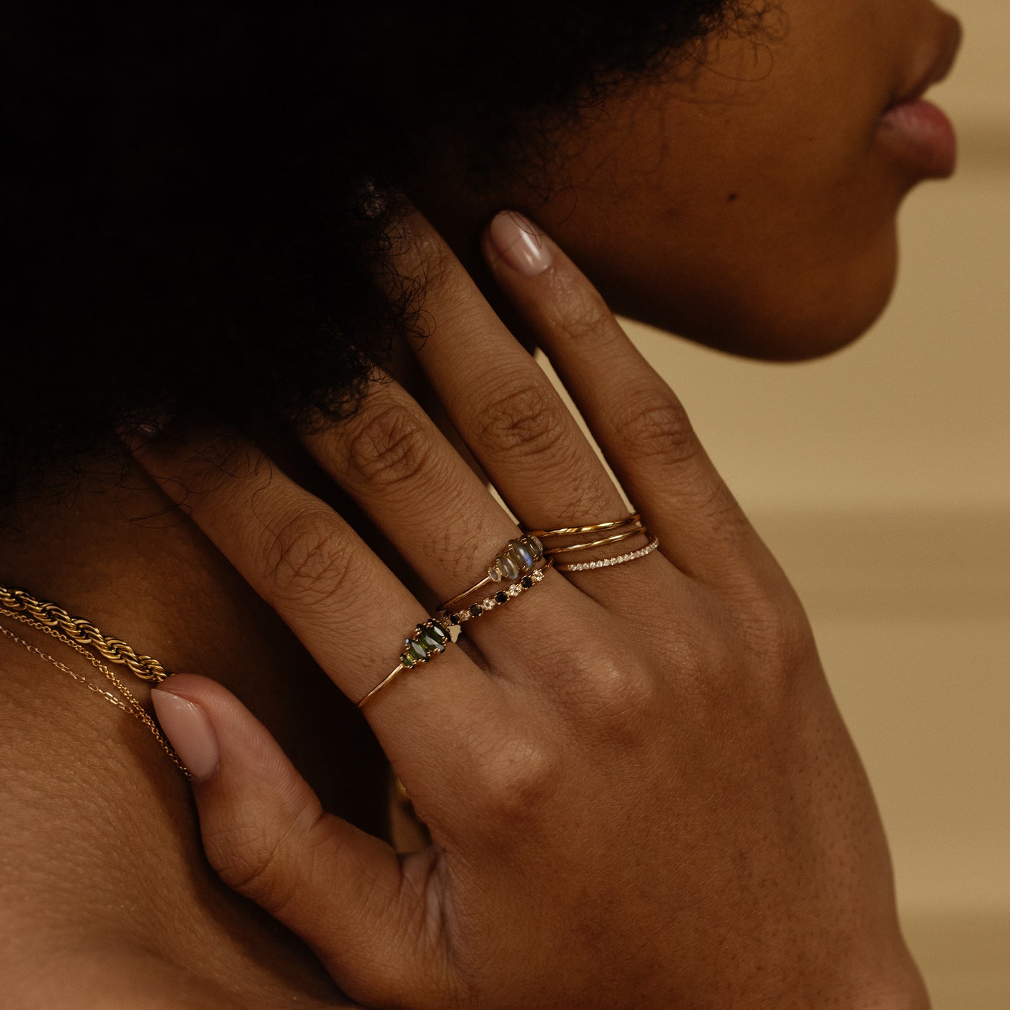 A close-up of a person’s hand with manicured nails wearing stacking rings, including the Renaissance Curved Stacking Ring in 14k yellow gold plate and cubic zirconia, gently touching their neck. The person also wears a gold chain necklace.