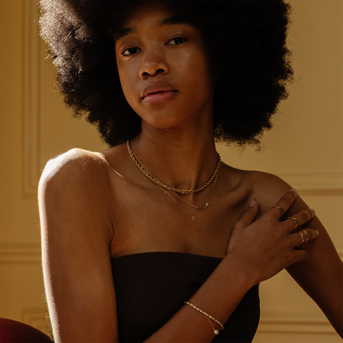 A woman with a natural afro wears gold layered necklaces, rings, and the Baguette Tennis Bracelet. She is dressed in a strapless black top and gazes calmly at the camera against a softly lit beige background.