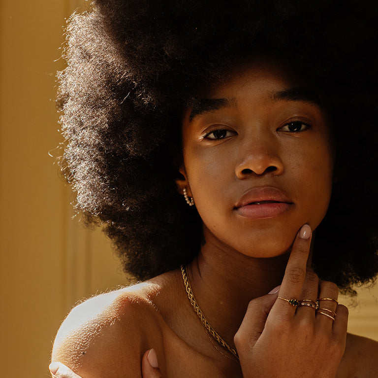 A young woman with a natural afro gazes at the camera, touching her chin. She wears Solid Gold Pearl Strand Hoops, bathed in warm sunlight against a soft yellow background.