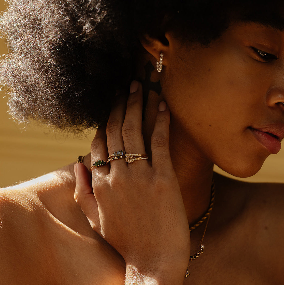 A woman with natural curly hair wears multiple rings, including the Solid Gold Essential Marquise Cluster Ring, along with a gold necklace and pearl earrings, as sunlight highlights her heirloom-quality jewelry and radiant skin.