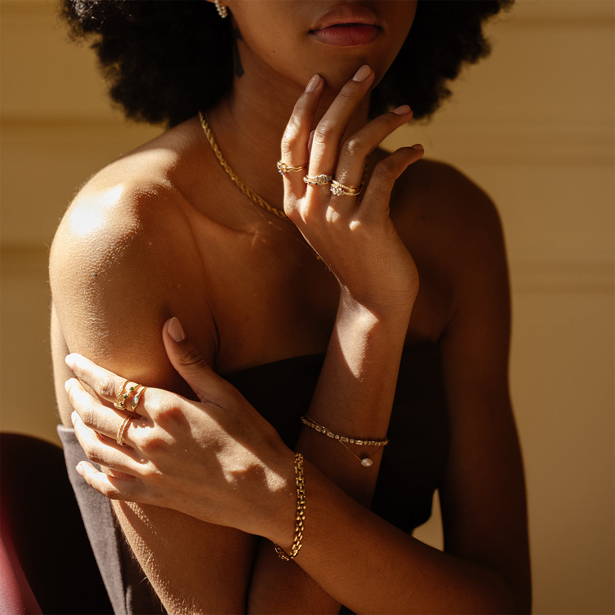 A woman with dark skin and curly hair wears a strapless black top and several accessories, including a Watch Band Bracelet, rings, a necklace, and earrings, posing with her hands elegantly near her face in warm light.
