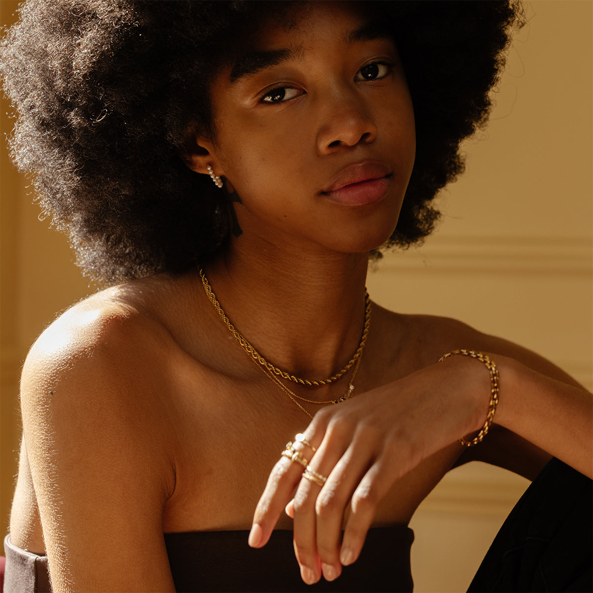 A young woman with an afro wears gold rings, a bracelet, earrings, and the Dreamworks Tennis Necklace with simulated colored diamonds. She gazes softly at the camera against a neutral, gently lit backdrop.