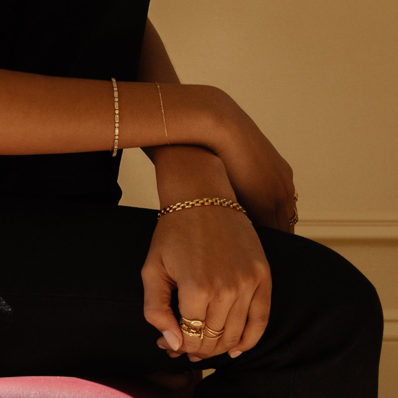 A person in black clothing sits with arms crossed, highlighting a Watch Band Bracelet alongside gold rings and a delicate gold bracelet against a neutral background.