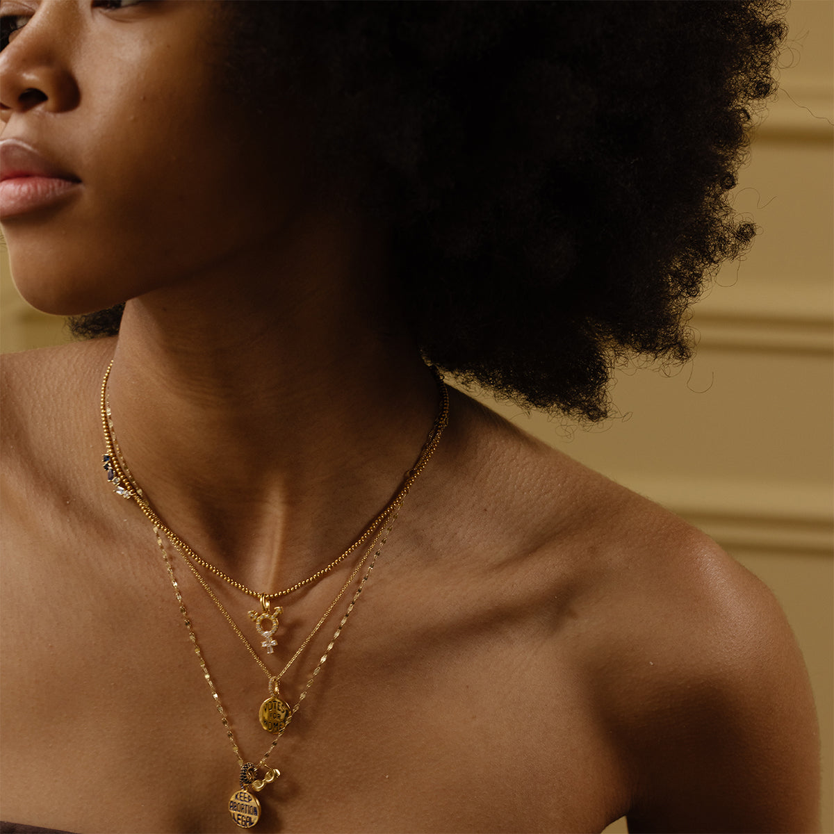 A woman with curly hair wears layered gold necklaces, including the Limited Edition Women's History Month Charm, and a strapless top, shown from the shoulders up against a beige background.