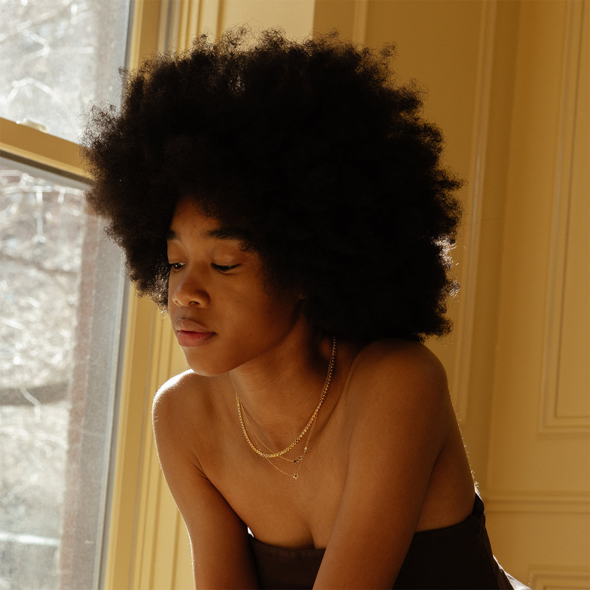 Wearing the shimmering Dreamworks Tennis Necklace, a person with a large afro and gold jewelry gazes thoughtfully downward while sitting by a sunlit window in a warmly lit room.