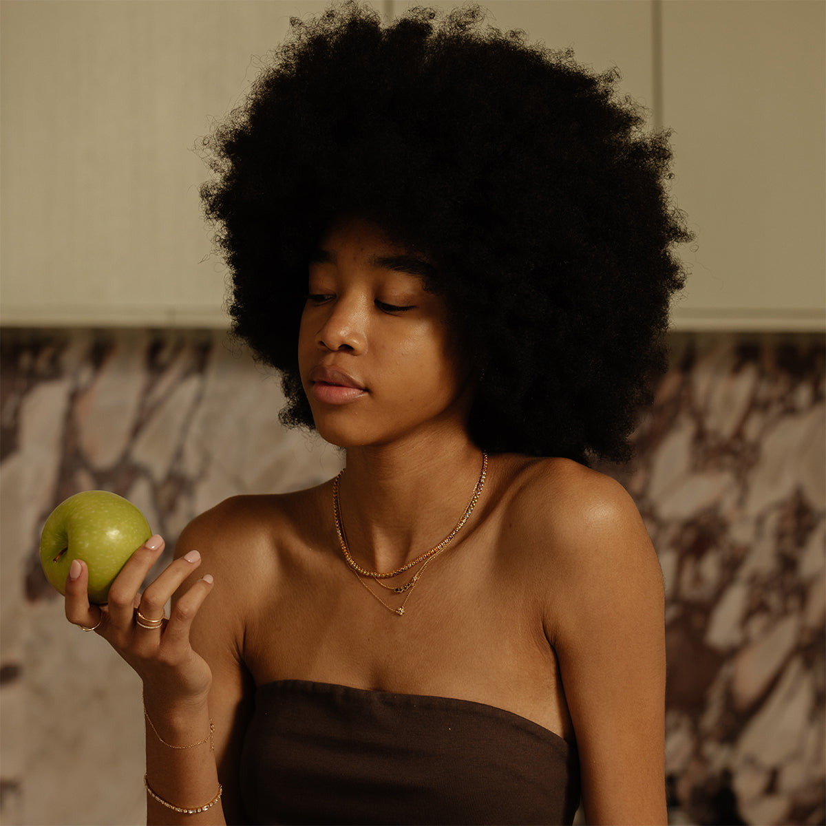 A young woman with a natural afro holds a green apple thoughtfully, wearing a brown strapless top and the Dreamworks Tennis Necklace, standing in a kitchen with marble-patterned walls.