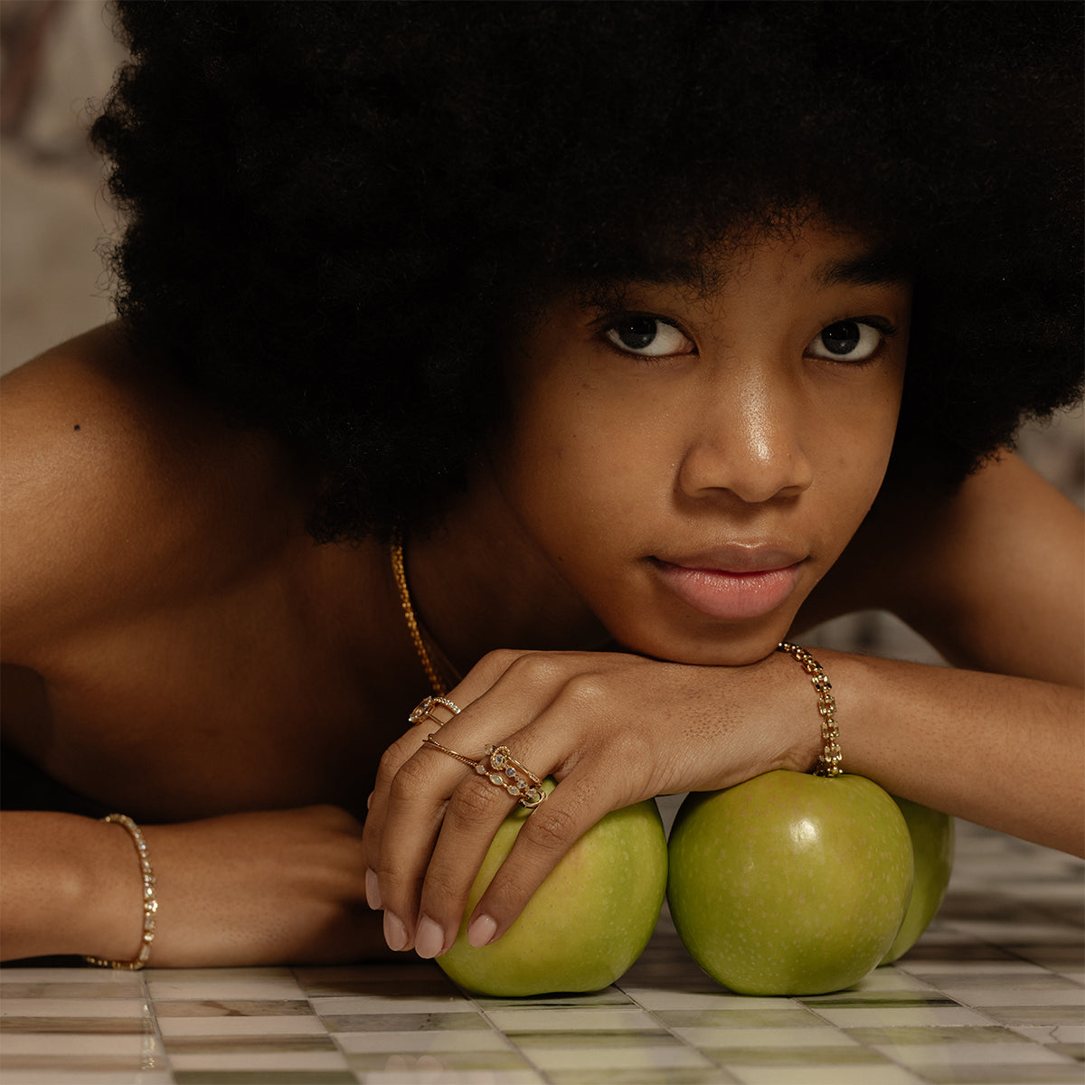 A young person with natural curly hair rests their chin on their hands, adorned with a Solid Gold Floating Gems Stacking Ring, placed on two green apples. They wear gold jewelry and gaze thoughtfully at the camera, on a tiled surface.