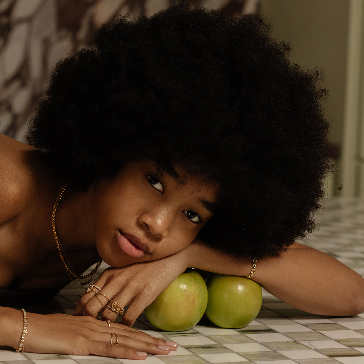 A woman with a natural afro rests her head on her arm above a marble surface, two green apples beneath her chin. She wears gold jewelry, including the Solid Gold Tourmaline Trio Stacking Ring, and gazes calmly at the camera.