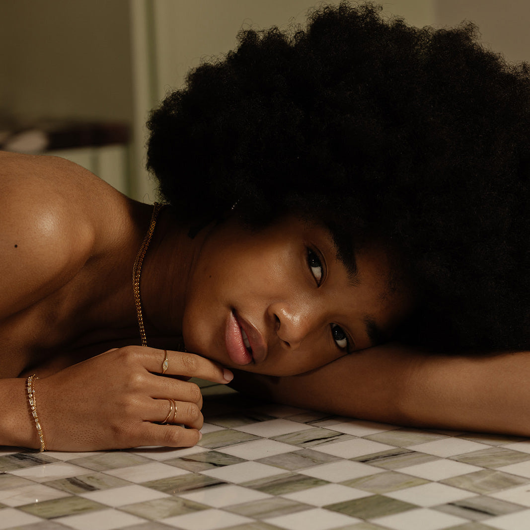 A woman with a natural afro hairstyle lies on a checkered marble surface, wearing delicate gold jewelry, including the Solid Gold Opal and Diamond Signet Ring on her finger, along with pieces around her wrist and neck.