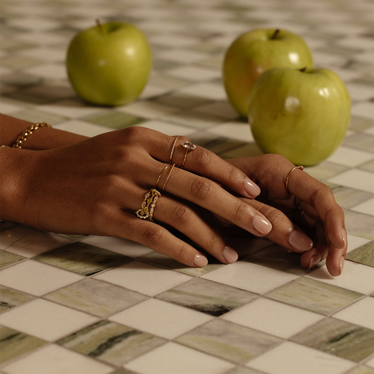 A pair of hands with manicured nails and multiple gold rings, including the Solid Gold Tourmaline Trio Stacking Ring, rests on a checkered countertop with three green apples in the background.