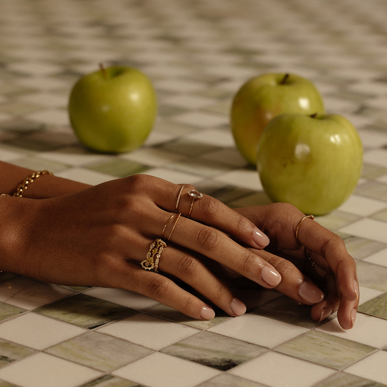 A close-up of manicured hands adorned with solid gold rings and bracelets, including the Solid Gold Gemstone Marquise Ring in White Topaz, resting on checkered tiles with three green apples in the background.