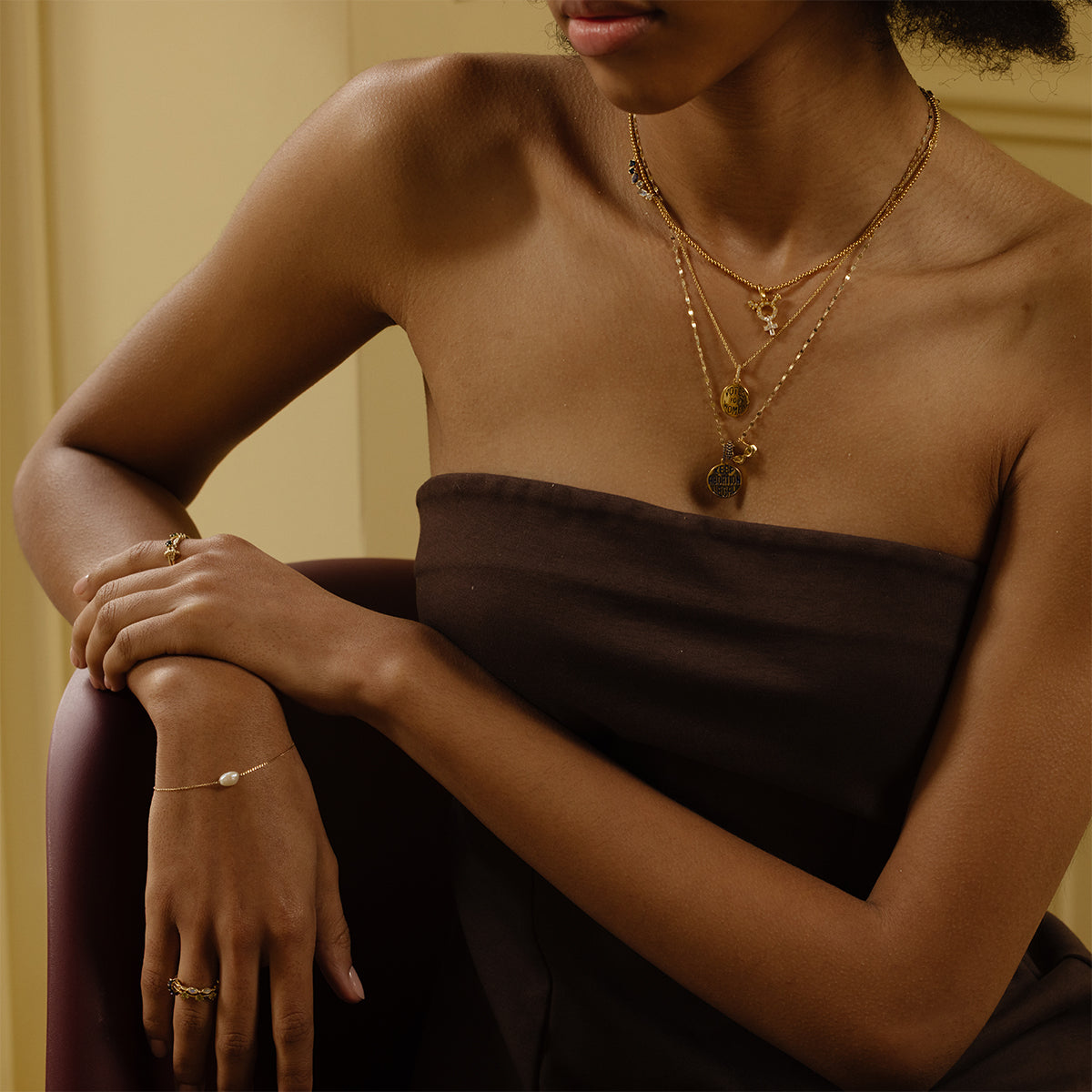 A woman in a brown strapless dress, layered gold necklaces, and a delicate bracelet with the Limited Edition Women's History Month Charm sits arms crossed against a softly lit beige background, highlighting unique women's jewelry.