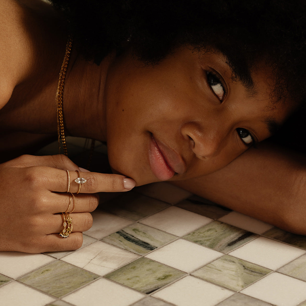 A woman with curly hair rests on white and green tiles, gazing at the camera. She wears solid gold rings, including the Solid Gold Gemstone Marquise Ring in White Topaz, and a gold necklace as her finger points at the tiles.