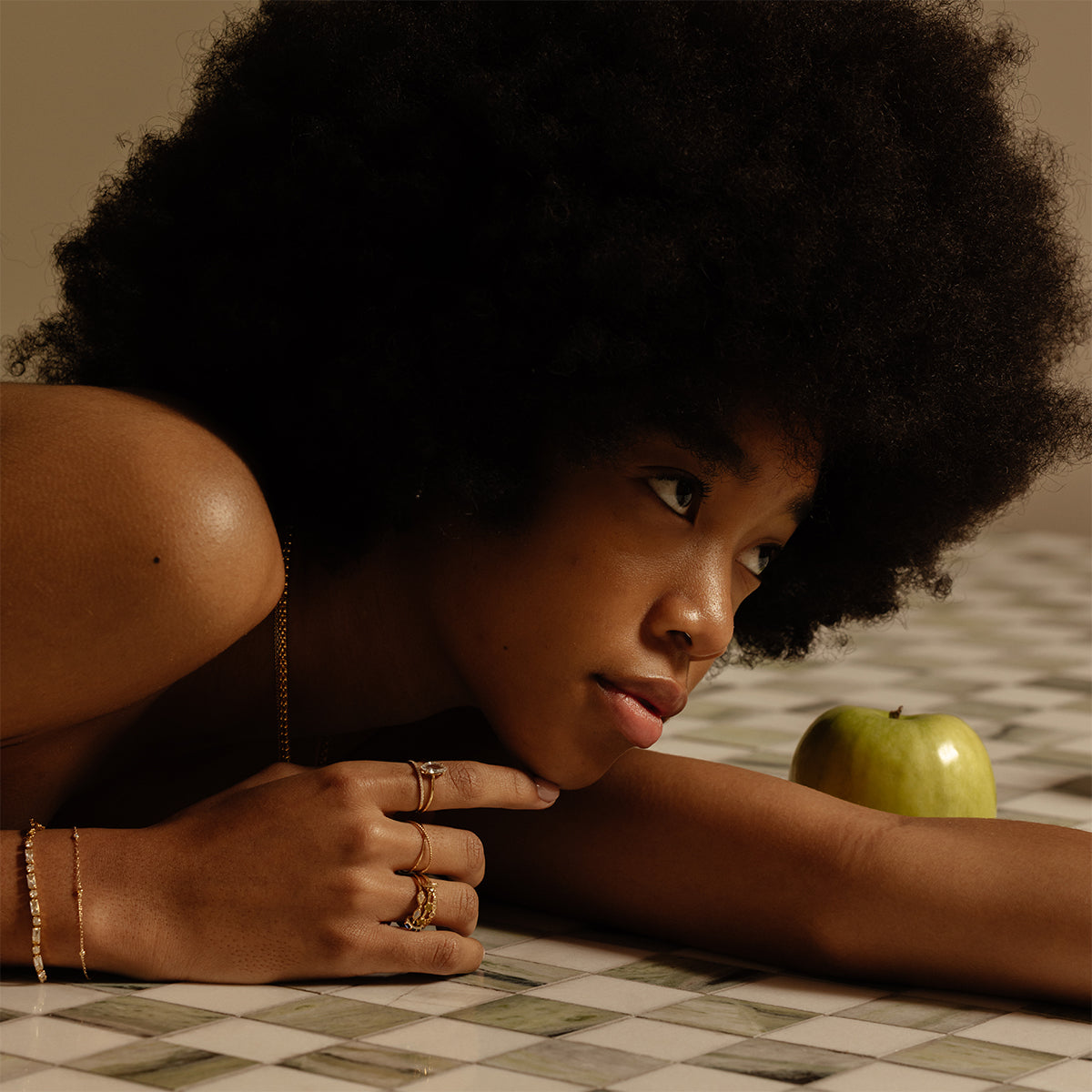 A woman with a natural afro lies on a tiled surface, gazing thoughtfully to the side. Her hand, adorned with the Solid Gold Tourmaline Trio Stacking Ring, touches her chin while a green apple rests near her arm.