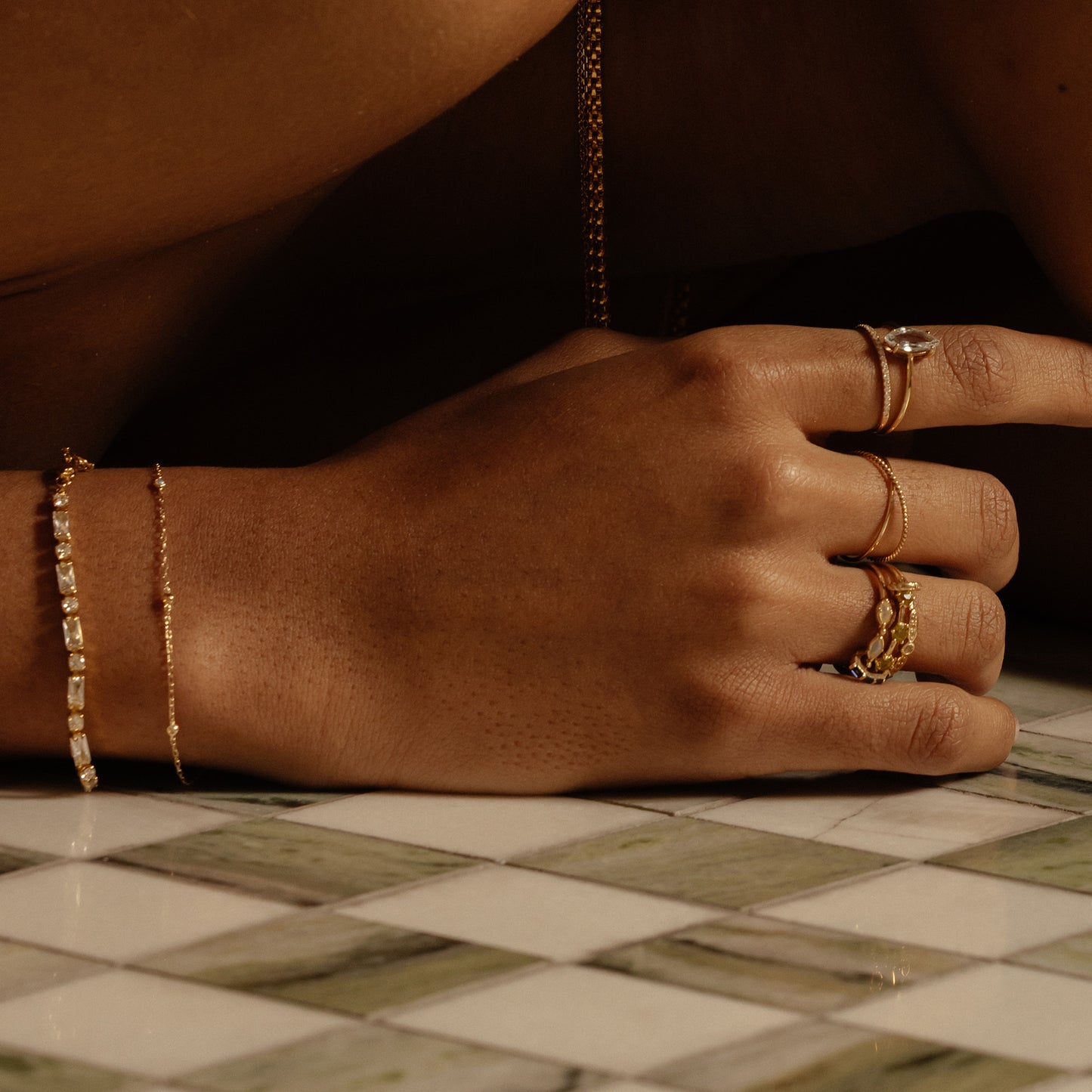 A close-up of a hand on green and white tiles, featuring multiple gold rings and the Solid Gold Everyday Opal Bracelet. Warm lighting accentuates the jewelry's shine and the skin's natural tones.
