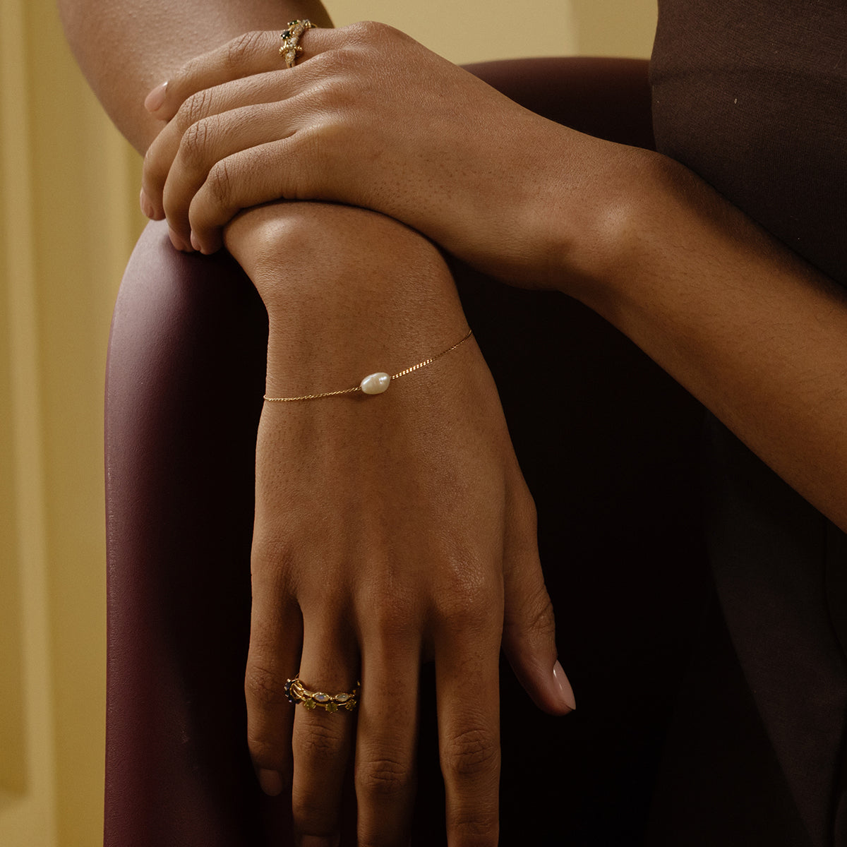 A close-up of a person in dark clothing resting their arm on a chair, wearing a delicate gold bracelet with a pearl and two Solid Gold Floating Gems Stacking Rings on their fingers.