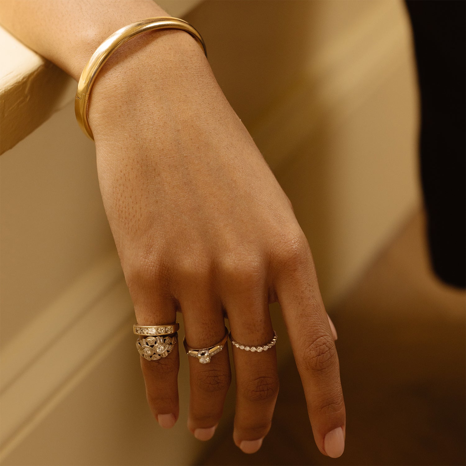 A close-up captures a hand on a ledge, adorned with several gold rings and the Vintage 9k Classic Bangle. The softly lit, neutral background highlights the elegant jewelry ensemble.