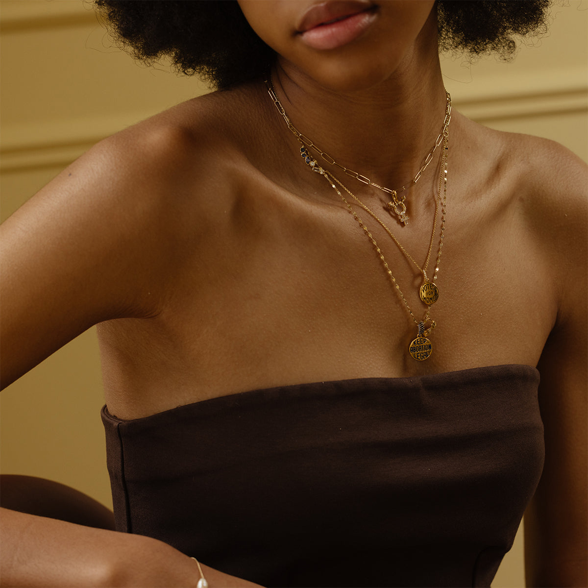 A woman with curly hair wears a dark brown, strapless top and layered gold necklaces featuring the Limited Edition Women's History Month Charm. She sits against a beige background, her face partially visible.