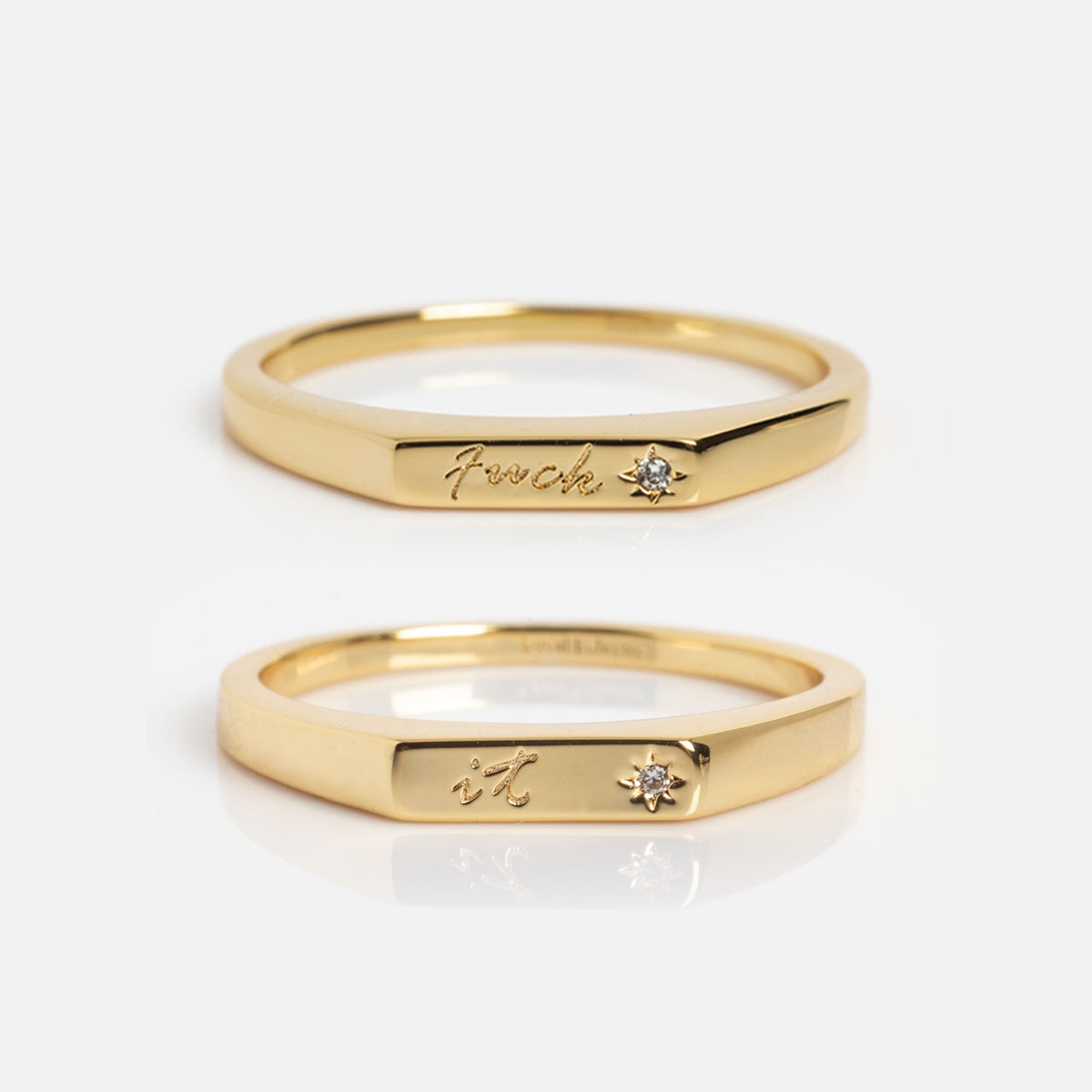 Two Tiny Conversation Stacking Rings with flat fronts, each engraved with scripted text and a small diamond—one reads "fuck" and the other "it." Both are shown on a plain white background.