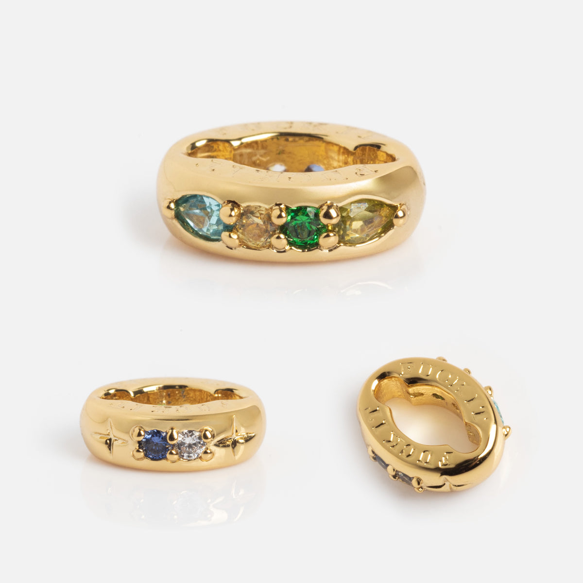 The Hidden Curse Word Spacer Charm is showcased on a white background. This chunky gold ring features small colored gemstones in unique arrangements and engraved side details.