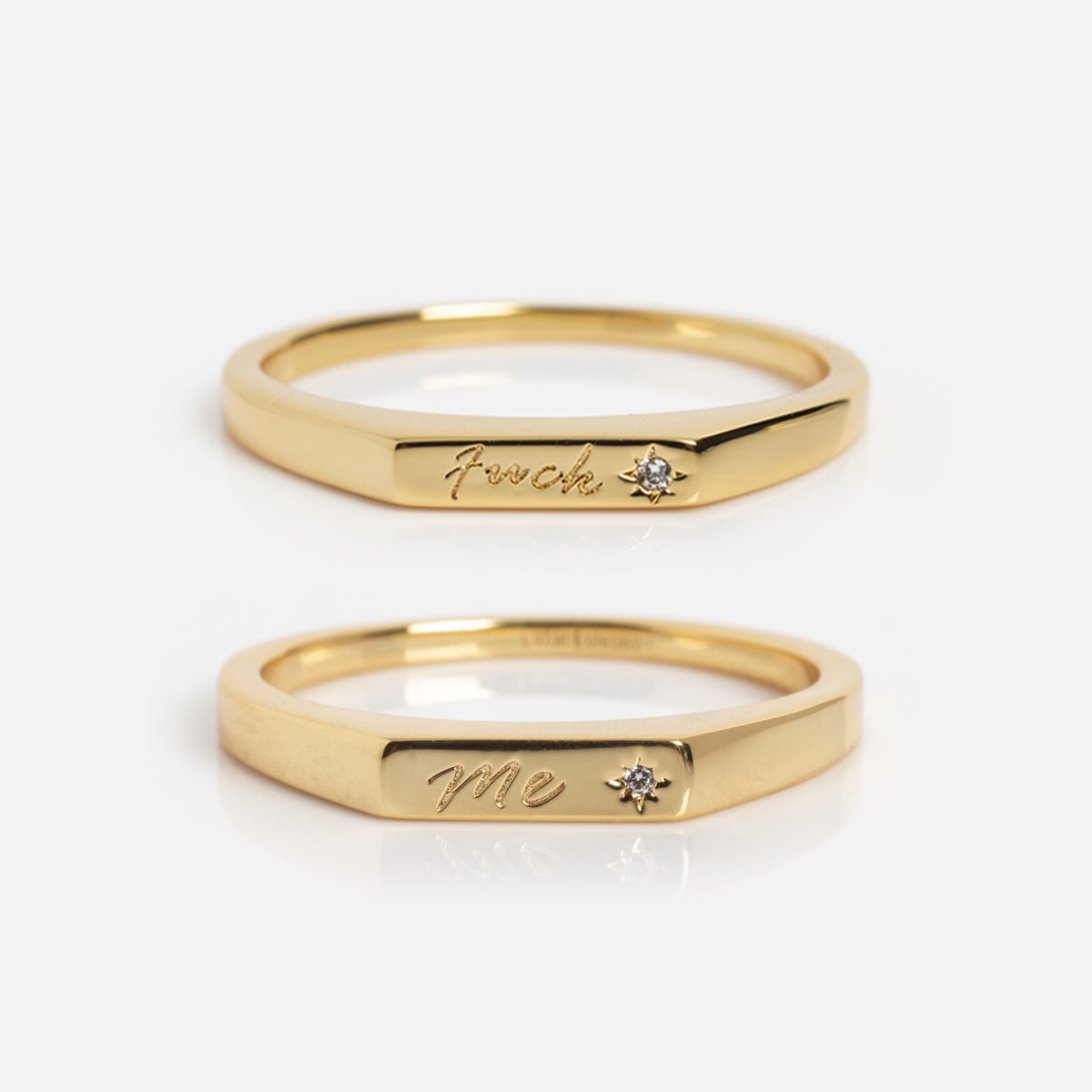 The Tiny Conversation Stacking Rings are two gold rings with flat fronts, each engraved with a word and set with a small diamond. One ring says "Fvck," the other "Me." Both are shown on a plain white background.
