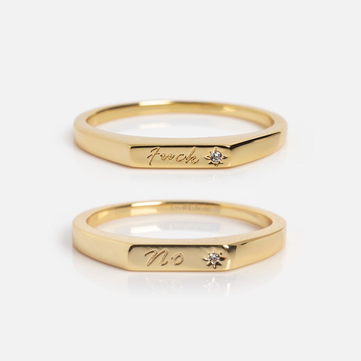 Two Tiny Conversation Stacking Rings in gold with small diamond accents—one engraved with "F*ck," the other "No." Their minimalist, modern design stands out against a plain white background.