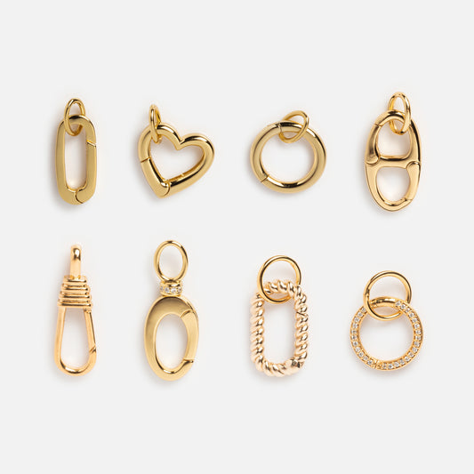 Eight gold jewelry charms in various shapes—oval, heart, circle, textured, and linked—are arranged in two rows on a light background. A Local Eclectic exclusive designed to pair perfectly with the Charm Clip.