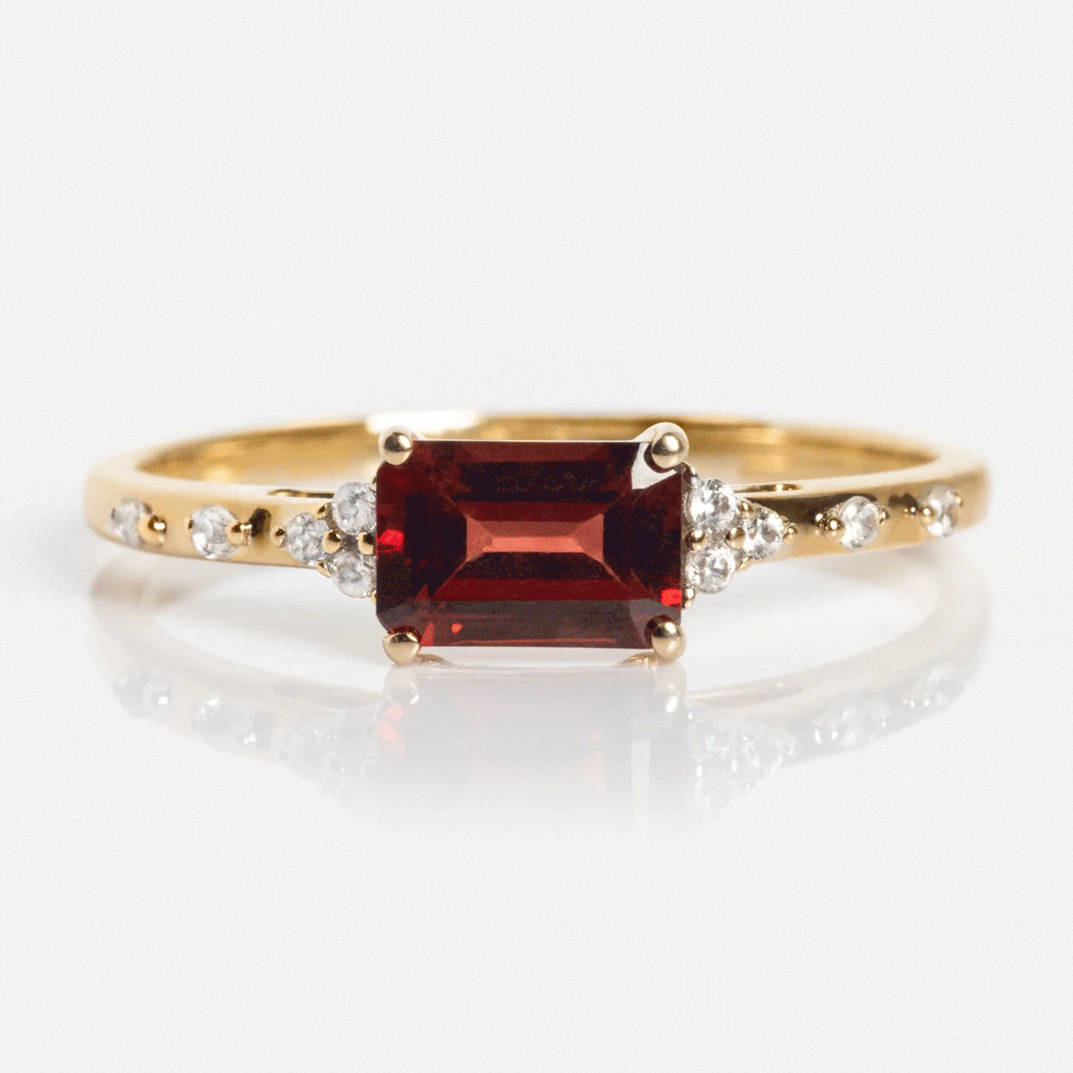 The Solid Gold Glimmering Birthstone Baguette Ring showcases a rectangular deep red center stone with small round white stones along its delicate band.