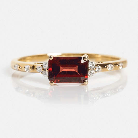 The Solid Gold Glimmering Birthstone Baguette Ring showcases a rectangular deep red center stone with small round white stones along its delicate band.