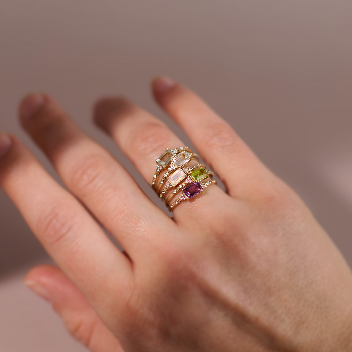 A hand showcases the Solid Gold Glimmering Birthstone Baguette Ring, set with green, pink, and clear stones—its radiant gold gleaming against a soft neutral backdrop, perfectly embodying Family Gold elegance.