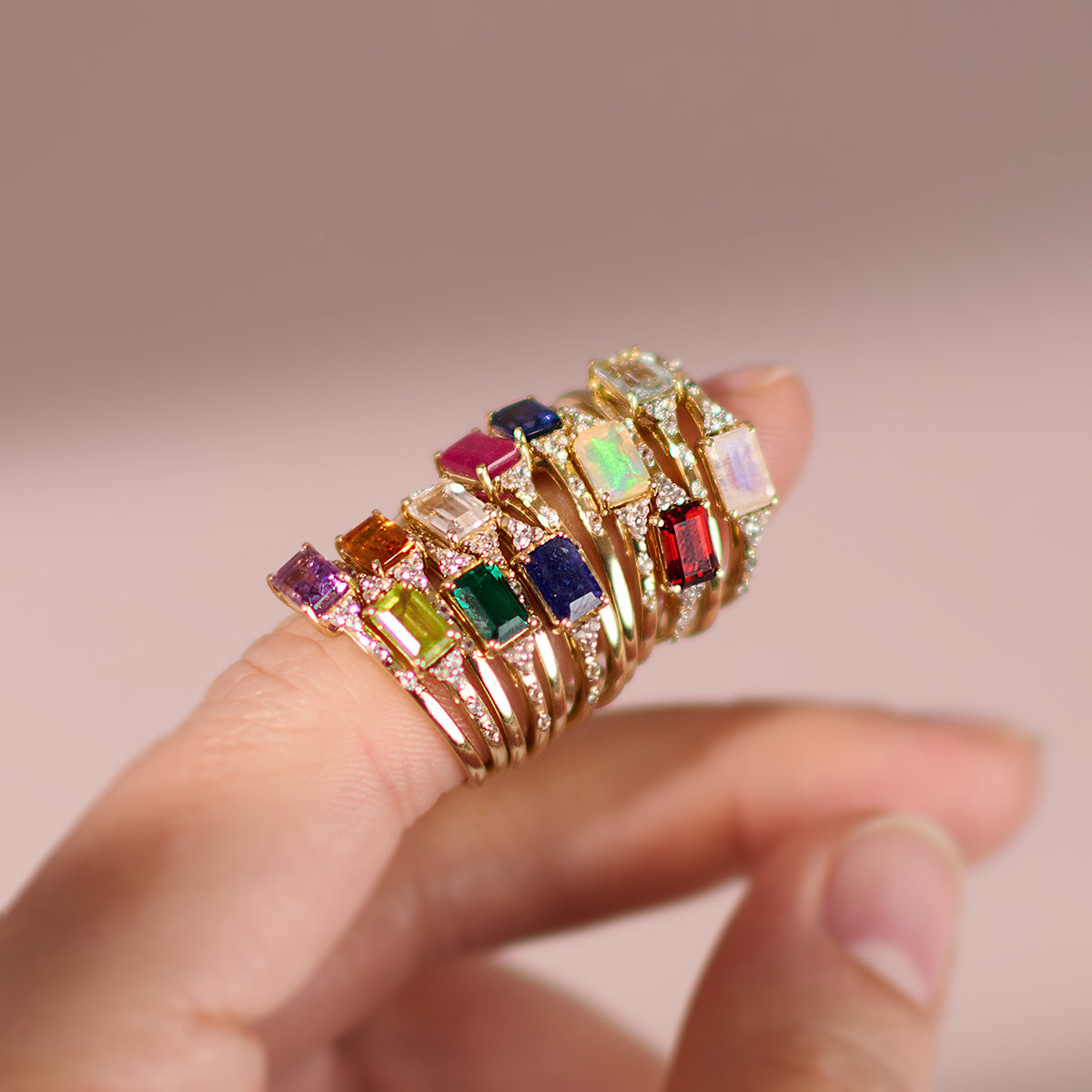A hand displays several Solid Gold Glimmering Birthstone Baguette Rings, each with a unique colorful gemstone, stacked on two fingers against a soft, light background.
