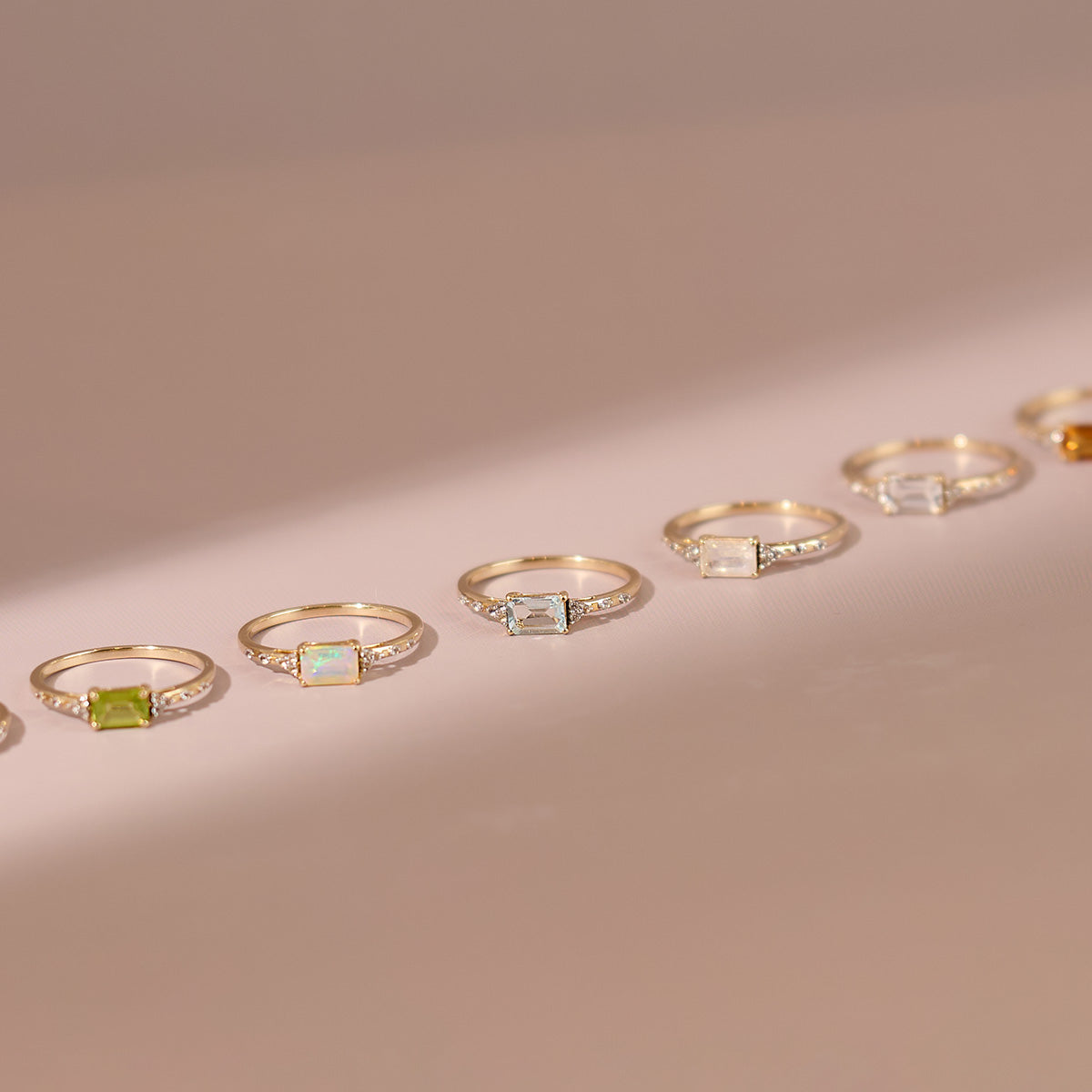 The Solid Gold Glimmering Birthstone Baguette Ring, featuring a sparkling rectangular gemstone, is displayed among gold rings with colorful gems on a light beige surface under soft lighting and gentle shadows.