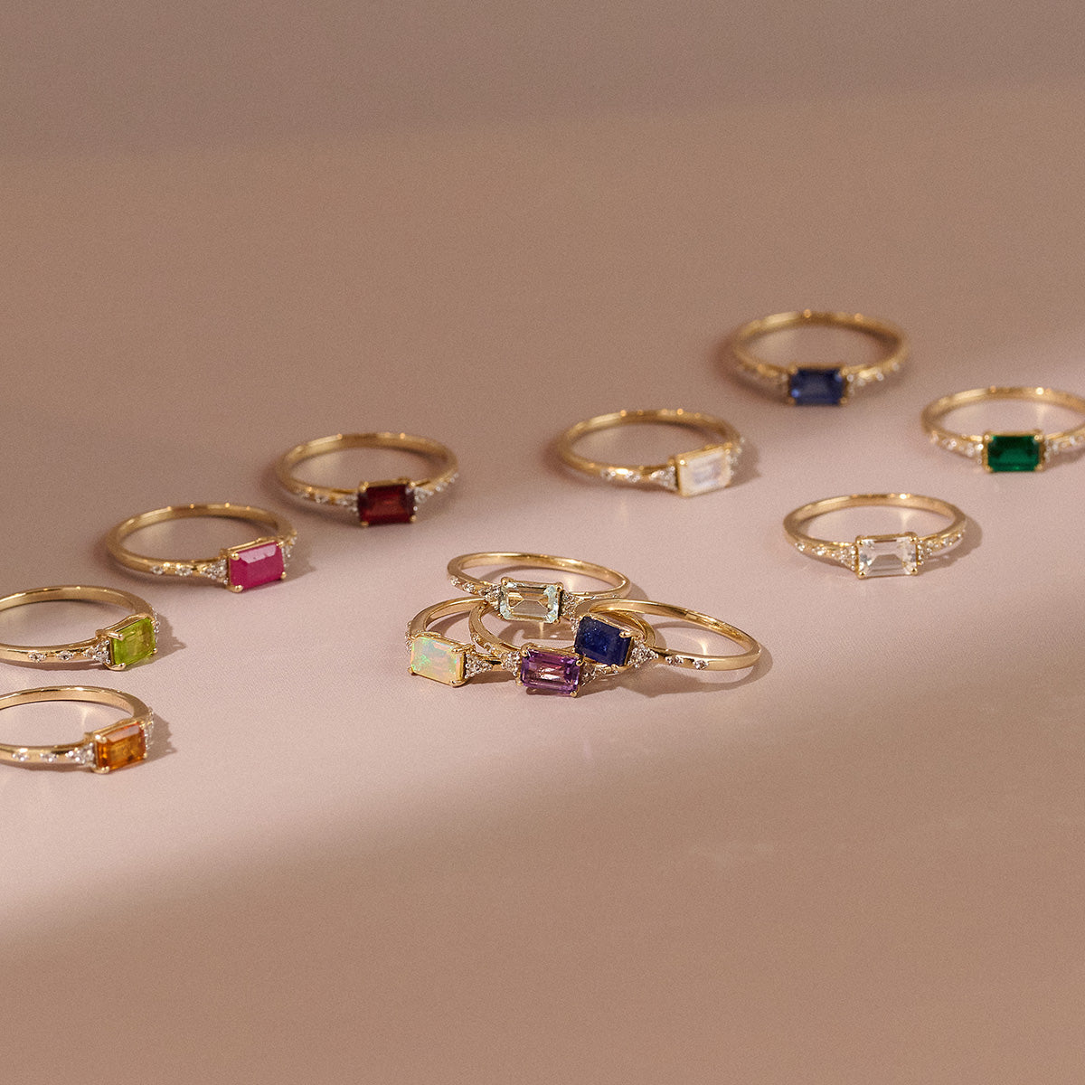 The Solid Gold Glimmering Birthstone Baguette Ring, featuring colorful gemstones and white topaz accents, is displayed on a light pink surface under soft lighting.
