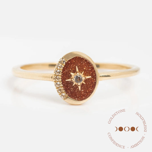 The Solid Gold Crystal Crescent Moon Ring for Manifestation features a crescent moon design with a sparkling centerpiece. Displayed on a white background with “Goldstone, Confidence, Ambition, Inspiration” in circular text.