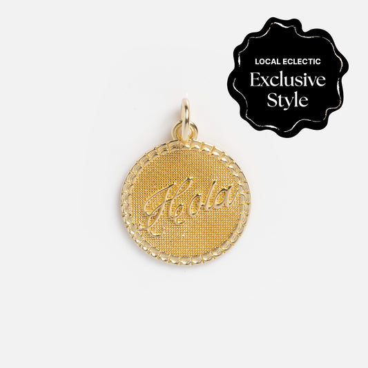 Inspired by the Zahava x Local Eclectic Globetrotter Charms, this yellow gold filled round pendant features Hola engraved in script with a textured background and decorative border—perfect travel-inspired jewelry.