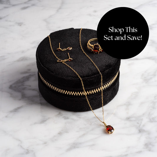 The Solid Gold 2025 January Capsule Garnet Full Set—necklace, earrings, and ring—is shown on a black velvet case with a gold zipper. A “Shop This Set and Save!” label appears over white marble. Includes a free gift.
