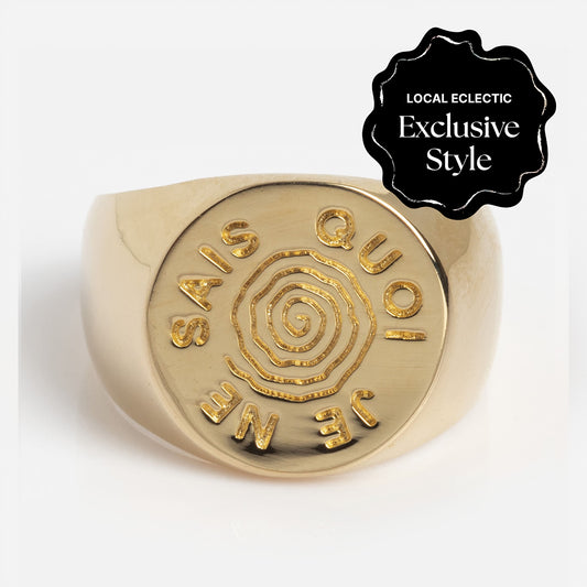 The I Like It Here Club x Local Eclectic Je Ne Sais Quoi Pinky Signet Ring features gold plating, a raised spiral design, and "JE NE SAIS QUOI" engraving; topped with a black badge marking it as a Local Eclectic Exclusive Style. Perfect for elevated everyday wear.