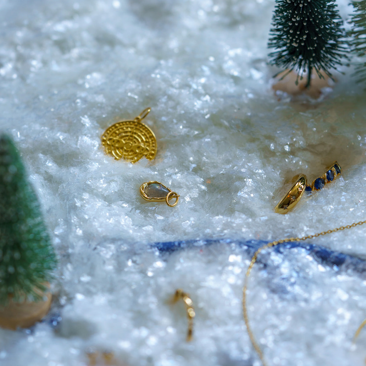 The 2025 Jewelry Advent Calendar features gold jewelry pieces—pendant, ring, and clasp—displayed on artificial snow with mini pine trees, evoking a festive winter atmosphere.