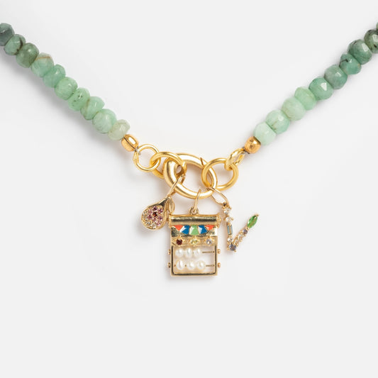 The Beaded Gemstone Charm Holder Necklace is a green beaded statement piece with a gold clasp, featuring three charms: a round pink stone, a rectangular charm with pearls and colorful stones, and a gold bar with a green gem.
