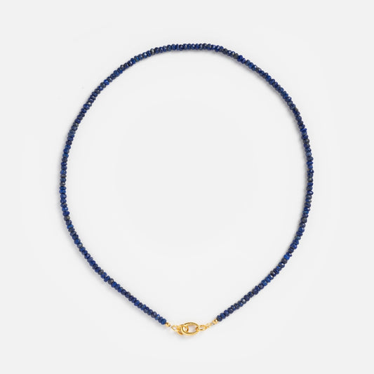 The Beaded Gemstone Charm Holder Necklace features small, round dark blue beads and a gold lobster clasp. This elegant, circular beaded necklace is displayed on a plain white background.