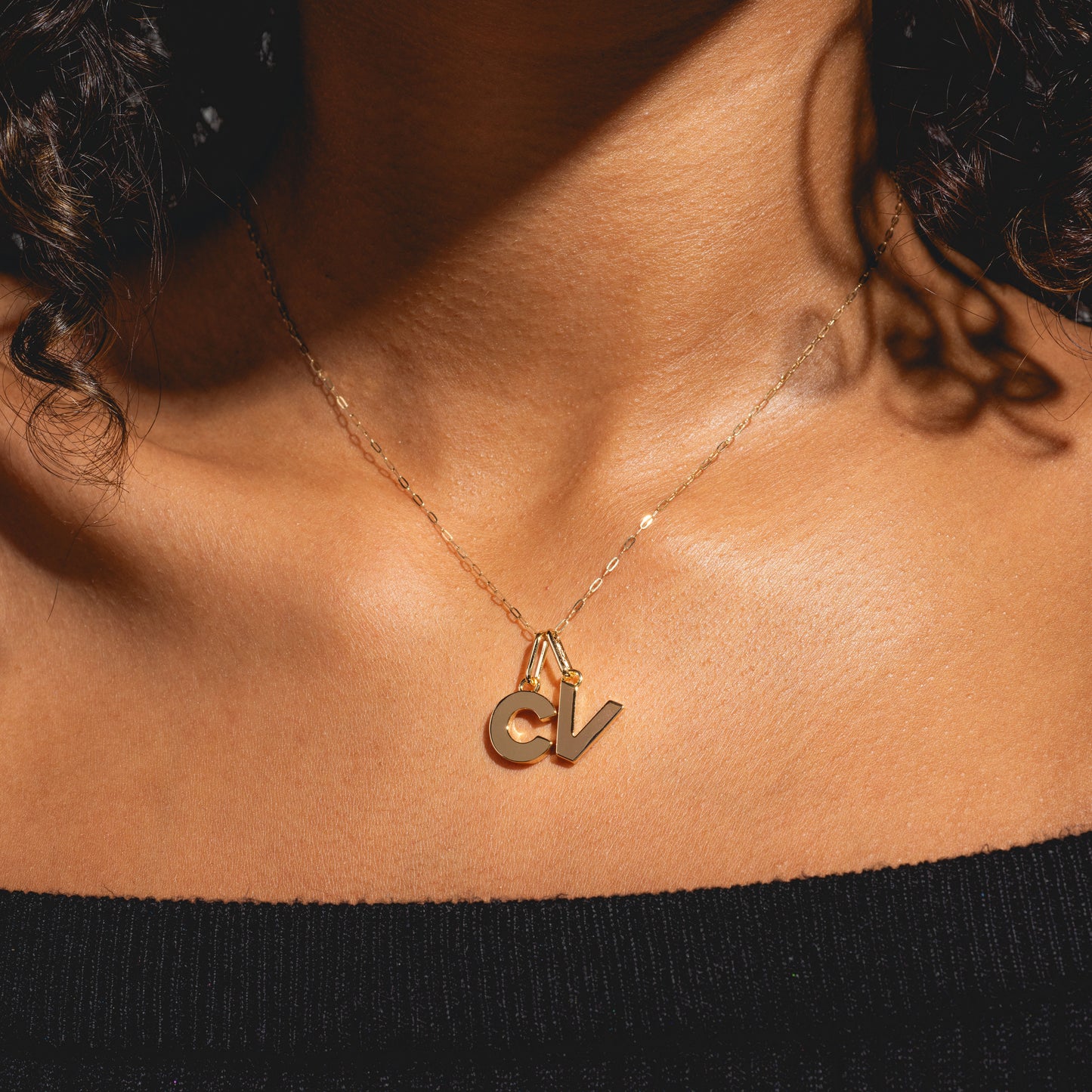 A woman wearing the Solid Gold Delicate Paperclip Chain with two large gold letter pendants, C and V, rests it against her bare upper chest and a black off-the-shoulder top.