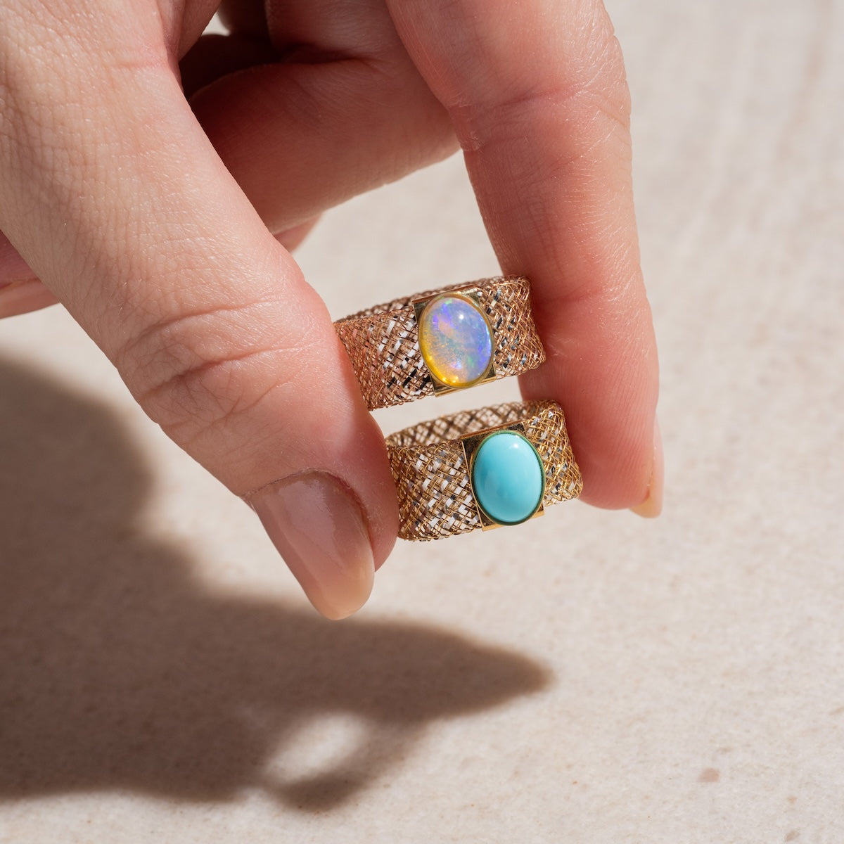 A hand holds two Solid Gold Mesh Gemstone Ring Sample Size 11, one set with an oval opal and the other with an oval turquoise, both on 9 mm yellow gold mesh bands against a neutral background.