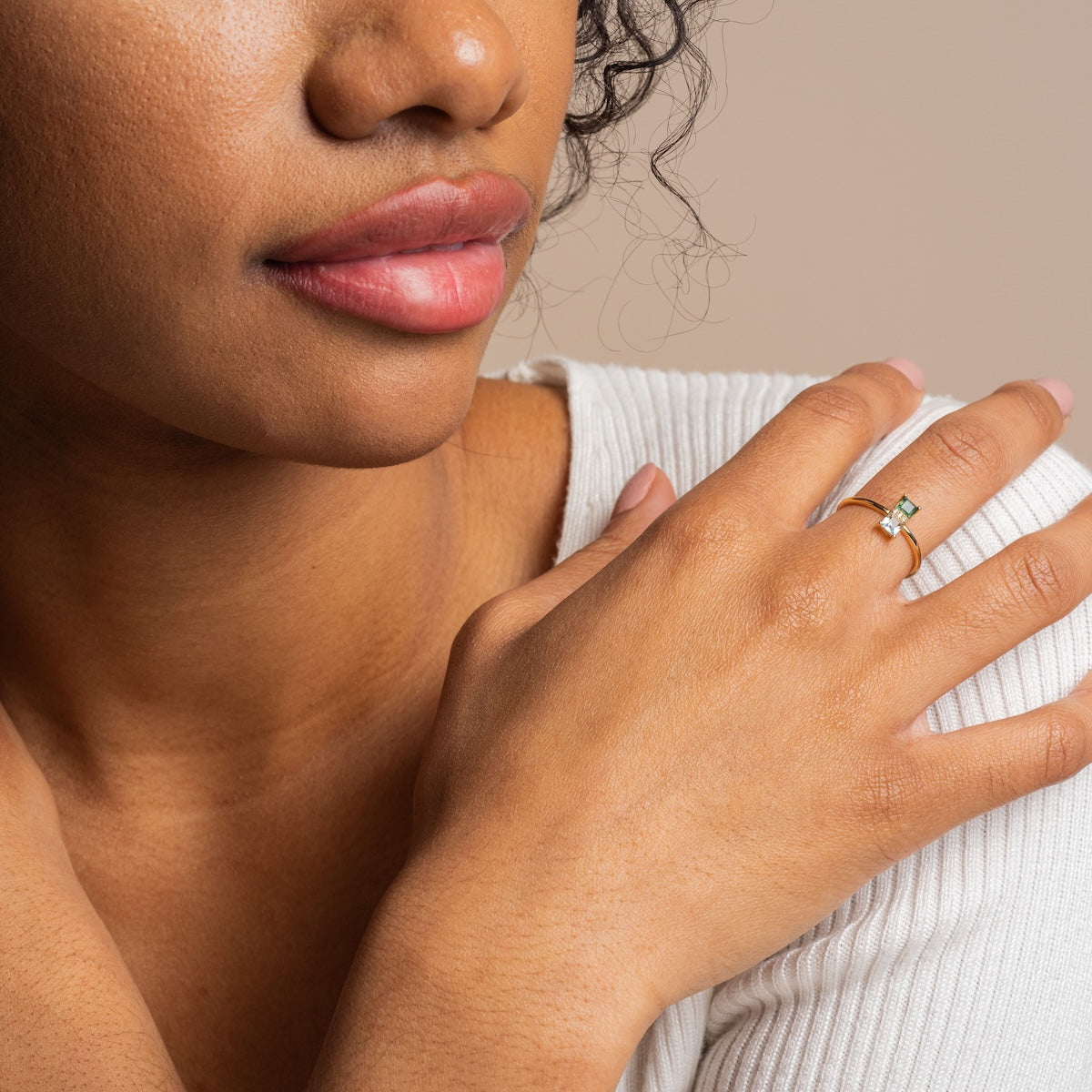 A woman in a white ribbed top rests her hand on her shoulder, showcasing the Solid Gold Toi et Moi North South Ring with a green gemstone. Her face is partially visible, drawing attention to her lips and curly hair.