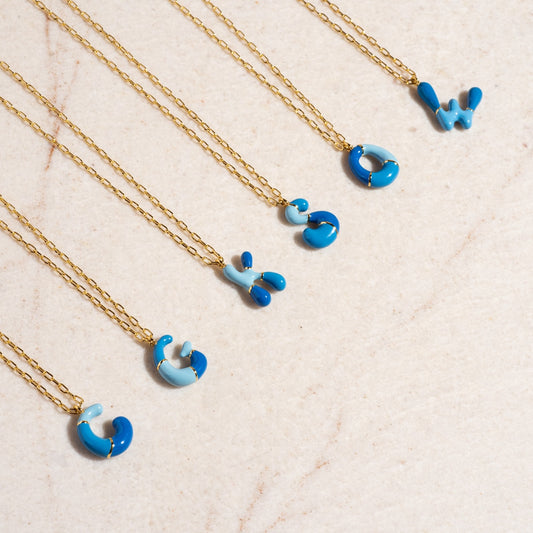 Six Blue Bubble Letter Necklace Sample pieces, each brass with 14k gold plating and blue/light blue enamel letter pendants (G, I, S, O, W, C), are arranged in a row on a light beige surface.