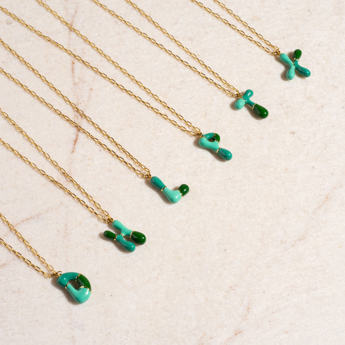 Six Green Bubble Letter Necklace Samples, each with a unique abstract teal and green enamel pendant on a 14k gold-plated brass chain, are displayed on a light beige textured surface.