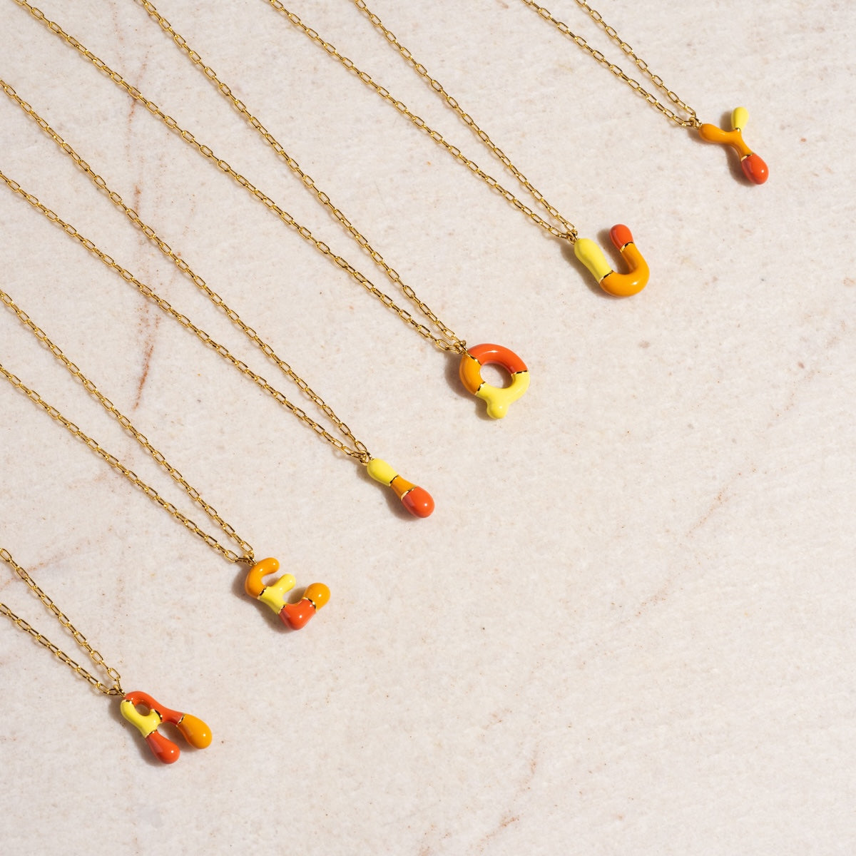 Six Orange Bubble Letter Necklace Samples, each featuring 14k gold plating with abstract yellow and orange enamel letter charms, are arranged diagonally on a light marble surface.